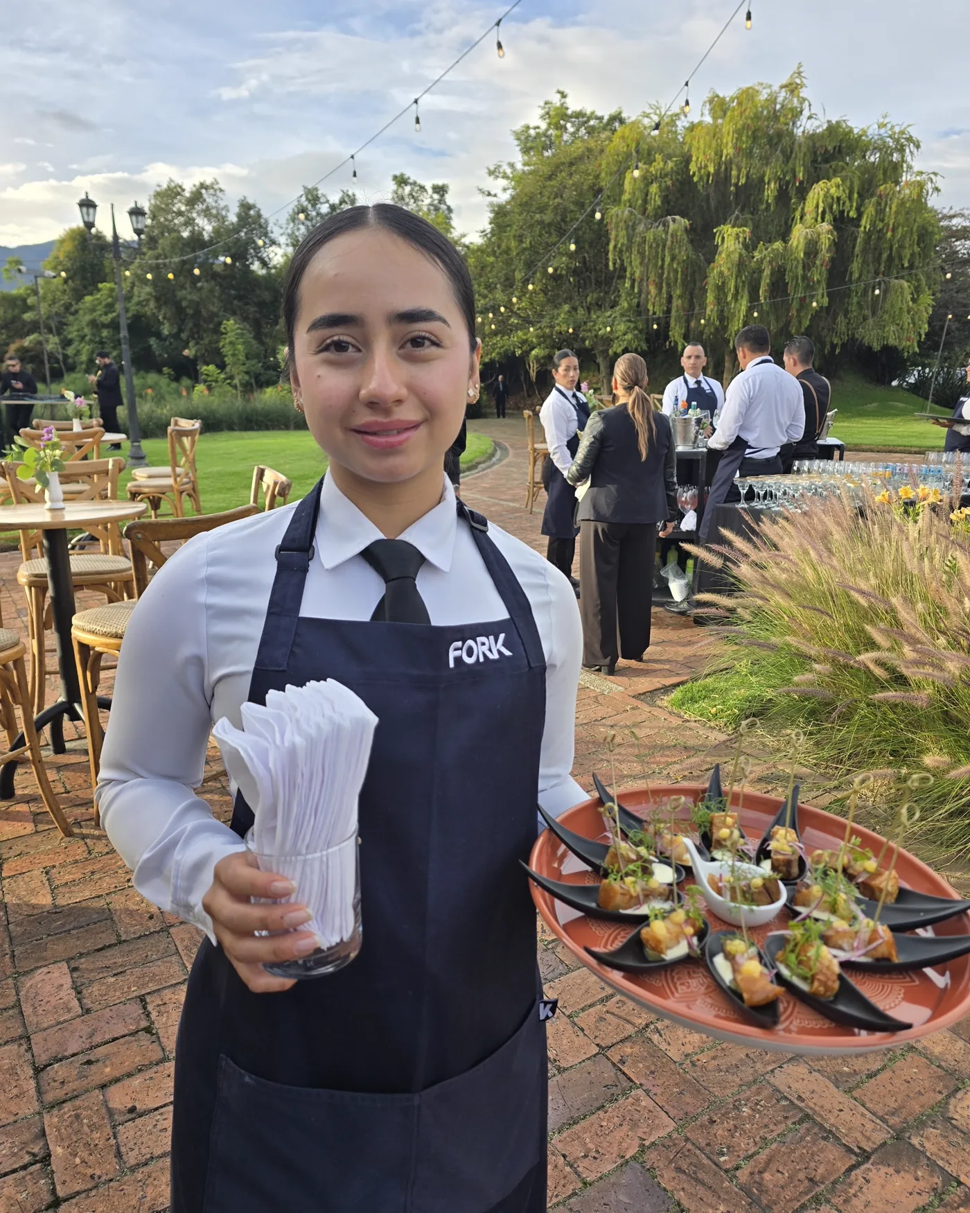 Mesera de Fork sonriendo con una bandeja de canapés en el jardín, al atardecer