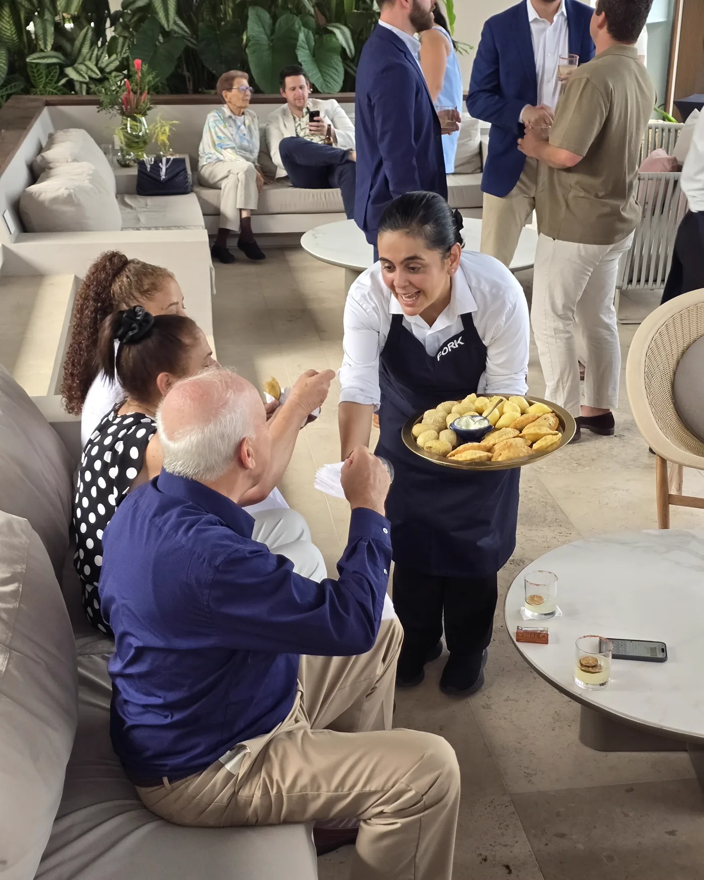 Una mesera acerca la bandeja a unos invitados sentados y uno de ellos estira la mano para servirse