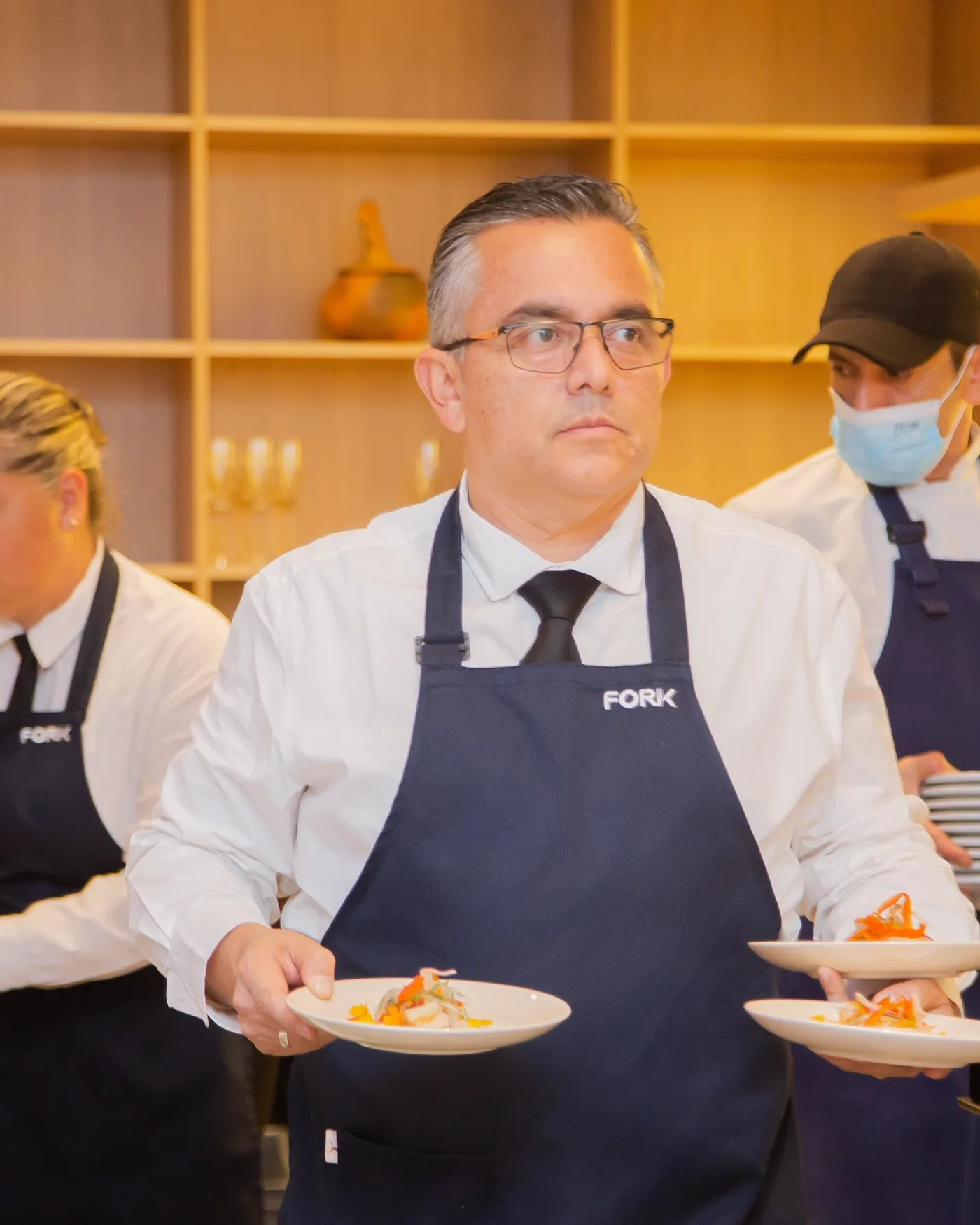 Maître de Fork saliendo con dos platos emplatados, con el equipo trabajando detrás