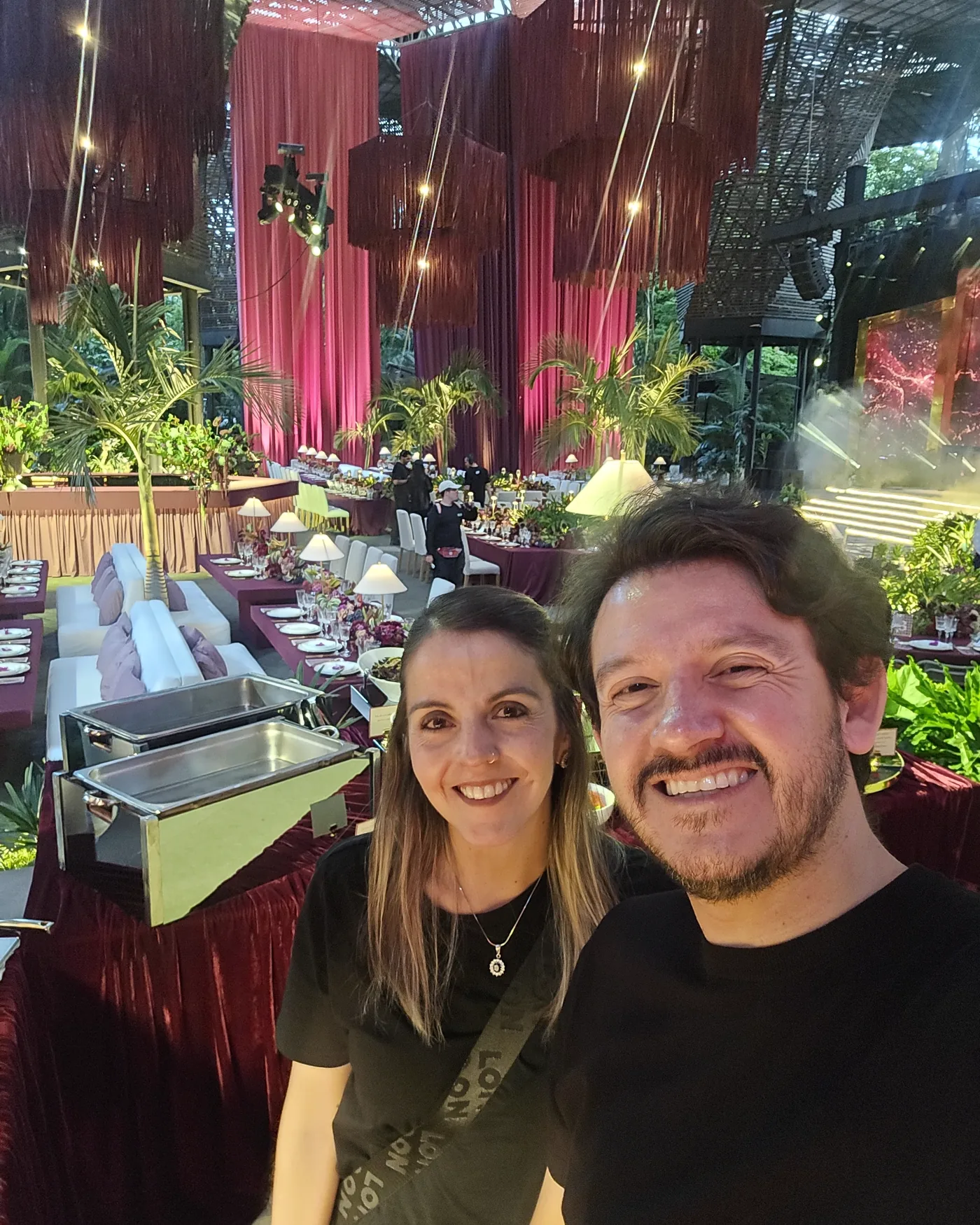 Camilo Alarcón y Carolina Ydrovo sonriendo en medio de un evento montado, con las mesas largas y la escenografía detrás